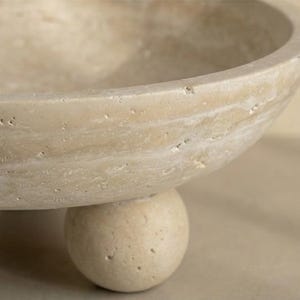 May include: A beige, stone bowl with a rounded shape, supported by a single, spherical base. The bowl has a smooth, curved rim and a textured surface. The neutral color and simple design suggest a decorative or functional item.