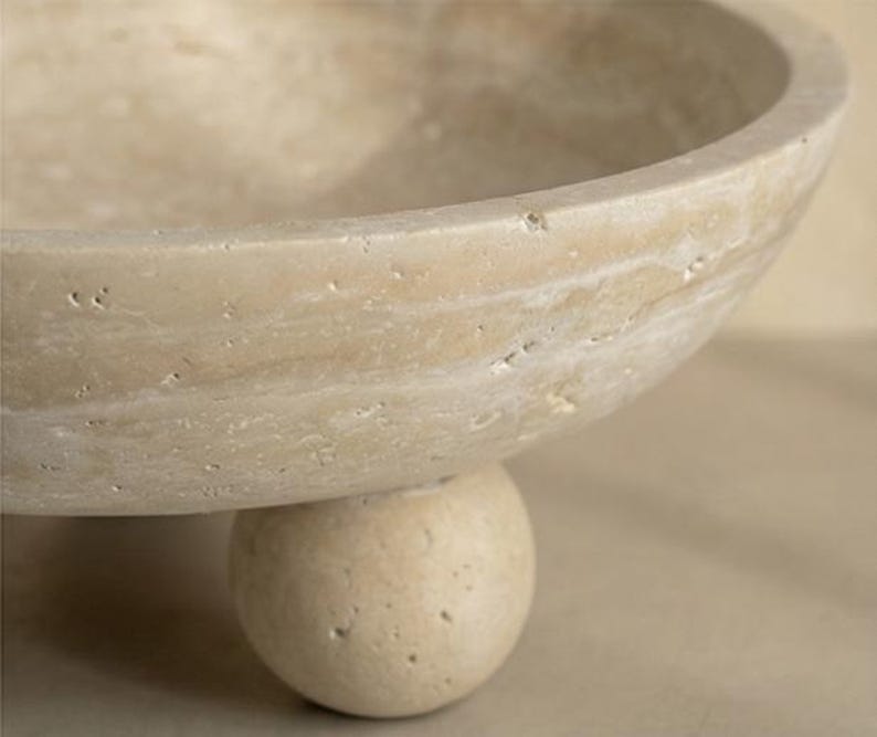 May include: A beige, stone bowl with a rounded shape, supported by a single, spherical base. The bowl has a smooth, curved rim and a textured surface. The neutral color and simple design suggest a decorative or functional item.