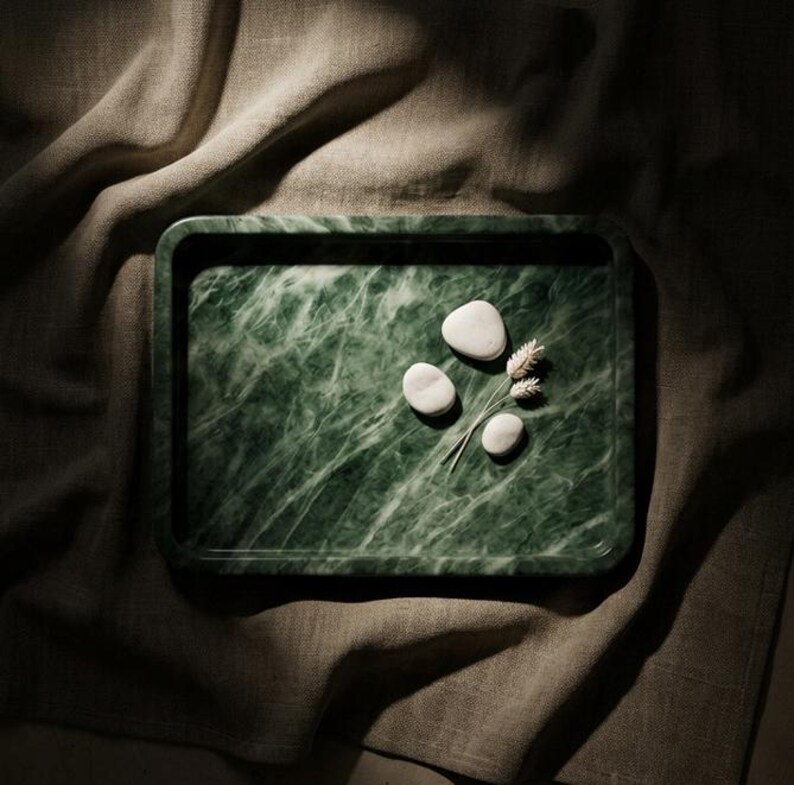 May include: A rectangular green marble tray with a white veined pattern. Three smooth, white stones and a small bundle of dried wheat are arranged on the tray. The tray is set on a neutral-coloured linen cloth.