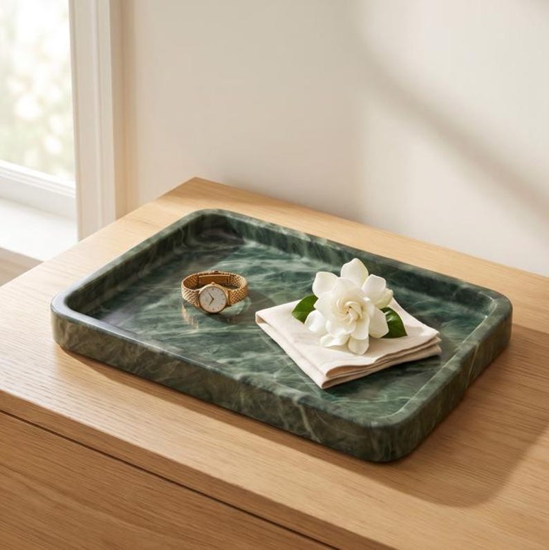 May include: A rectangular green marble tray with rounded corners. A gold wristwatch with a mesh strap and a white face sits on the tray, alongside a folded white napkin and a white flower.
