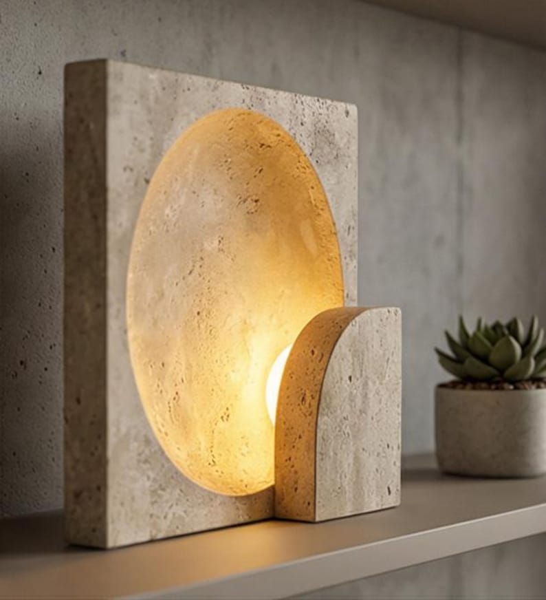 May include: A modern, rectangular stone lamp with a semi-circular cutout that emits warm light. The lamp has a matching stone base and is placed on a shelf. A small potted succulent is visible in the background.