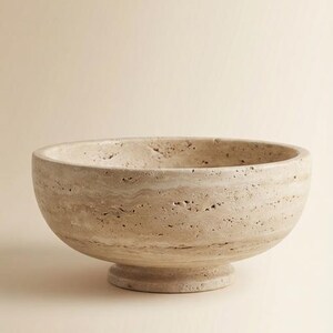 May include: A beige stone bowl with a rounded shape and a pedestal base. The bowl has a textured surface with natural variations in colour and pattern. This decorative bowl is suitable for home decor.