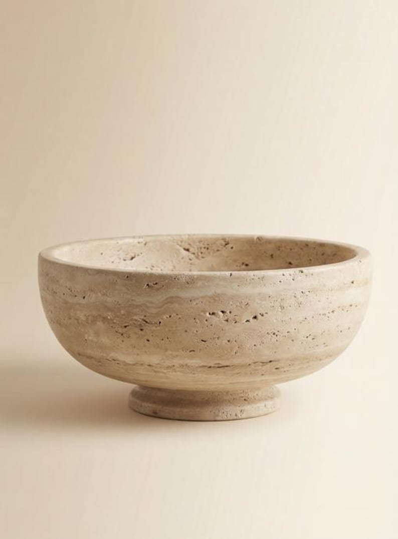 May include: A beige stone bowl with a rounded shape and a pedestal base. The bowl has a textured surface with natural variations in colour and pattern. This decorative bowl is suitable for home decor.