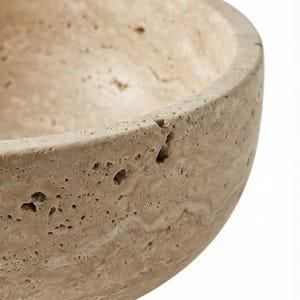 May include: Close-up of a beige stone bowl with a rounded shape. The bowl has a rough, textured surface with small holes and natural colour variations. Likely made of travertine or similar stone.