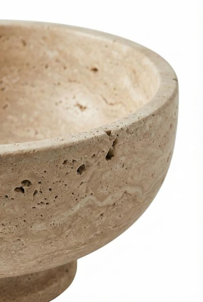 May include: Close-up of a beige stone bowl with a rounded shape. The bowl has a rough, textured surface with small holes and natural colour variations. Likely made of travertine or similar stone.