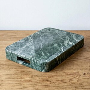 May include: A rectangular serving tray made of green marble with white veining. The tray has rounded corners and a rectangular handle cut-out on one side. The tray is displayed on a wooden surface.