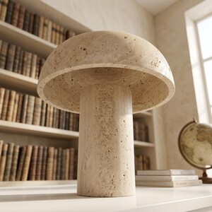 May include: A mushroom-shaped table lamp crafted from beige, porous travertine stone. The lamp features a wide, rounded dome shade resting atop a cylindrical base, showcasing the natural, textured, and pitted surface of the stone material.