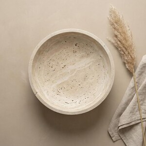 May include: A round, beige stone bowl with a speckled pattern. The bowl is empty and sits on a neutral-toned surface. A dried plant and a linen cloth are next to the bowl, creating a minimalist aesthetic.