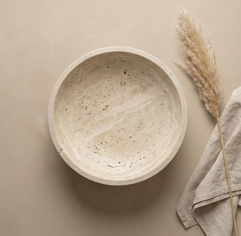 May include: A round, beige stone bowl with a speckled pattern. The bowl is empty and sits on a neutral-toned surface. A dried plant and a linen cloth are next to the bowl, creating a minimalist aesthetic.