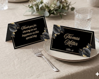 Könnte beinhalten: Zwei schwarze, gefaltete Tischkarten mit goldenen Rändern und goldenen Blattmotiven. Eine Karte trägt den Text "Thank you for sharing in our happiness on this special day", die andere "Thomas Miller" mit "Table-12" in goldener Schreibschrift.
