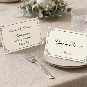 Può includere: Due segnaposto in carta piegata color crema con testo in inchiostro nero e bordi floreali decorativi agli angoli. Un cartoncino riporta Amelia and Damian Thank you for joining us on our happy day. L'altro cartoncino riporta Charlie Brown Table 15.