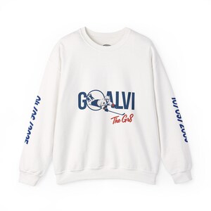 Op de afbeelding: Witte longsleeve sweatshirt met blauwe en rode tekst die "GOALVI The Gr8" luidt en een afbeelding van een hockeyer. De mouwen hebben blauwe tekst die "All In The Game" en "The Gr8" luidt