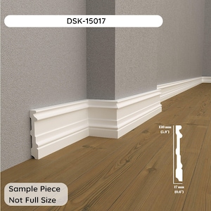 Op de afbeelding: Een witte plint met een decoratief profiel geïnstalleerd langs een hoek waar grijze muren een bruine houten vloer ontmoeten. Een diagram toont de profielhoogte van 150 mm en dikte van 17 mm. De tekst luidt DSK-15017 en Sample Piece Not Full Size.