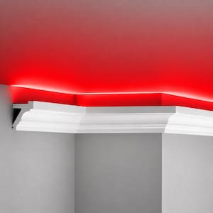 May include: A white crown moulding profile installed along the top of a grey wall, featuring a recessed channel that emits a bright, continuous red light glow against the ceiling, creating a sharp contrast between the white trim and red light.