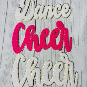 May include: Three script-style patches featuring the words Dance, Cheer, and Cheer. The top and bottom patches are white with gold trim, while the middle patch is bright neon pink with gold trim. Each patch has a textured, fuzzy surface finish.