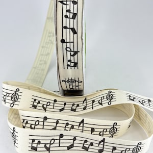 May include: A roll of cream-coloured ribbon printed with a repeating pattern of black musical notes and staves. The ribbon is unrolled, displaying the musical notation. Suitable for crafts and gift wrapping.