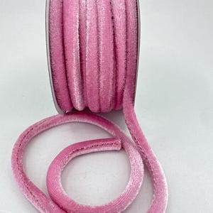 May include: A roll of pink velvet piping ribbon. The ribbon is a soft, plush material with a rounded shape. The roll is partially unwound, showing the ribbon's texture and colour. The background is a plain white surface.