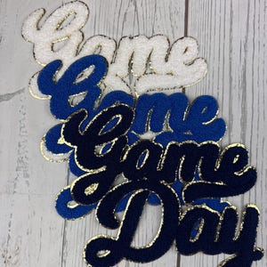 May include: A set of four layered chenille patches featuring the text Game Day in a cursive font. The patches are stacked, with the top layer in white, the middle layers in royal blue, and the bottom layer in navy blue, all with gold glitter trim.