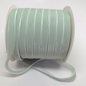 Puede incluir: Una bobina de cinta de terciopelo verde claro. La cinta está enrollada alrededor de una bobina de cartón blanco.