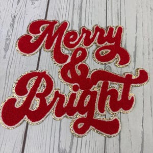 May include: A red chenille patch with gold glitter trim featuring the text Merry & Bright in a cursive script font. The patch has a textured surface and a layered appearance with the red lettering outlined by a sparkling gold border.