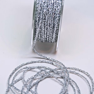 May include: A spool of silver glitter ribbon. The ribbon is wound around a clear plastic spool and is partially unwound, showing the texture of the ribbon.