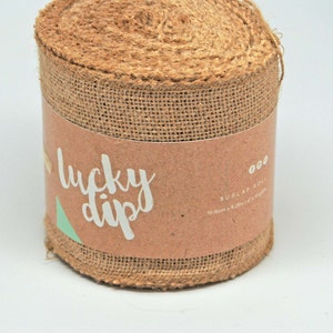 May include: A roll of burlap ribbon with a brown paper label. The label reads "lucky dips" in white script with a green triangle. The burlap roll measures 10.36m x 9.25in.