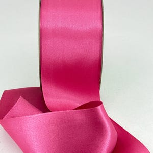 May include: A roll of vibrant fuchsia satin ribbon. The ribbon is unwound, revealing a smooth, glossy texture. The color is a bright, saturated pink, perfect for crafting, gift wrapping, or decorative purposes. The ribbon is likely made of polyester.