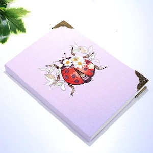 Pode incluir: Um diário de cor lavanda com um design de joaninha bordada detalhada. A joaninha é vermelha com pintas pretas e flores brancas. O diário tem protetores de canto de latão.