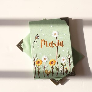 May include: A light green embroidered journal with the name "Maria" in gold thread. The cover features a floral design with yellow and white flowers, a bee, and decorative corner protectors.