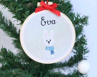 Può includere: Ornamento natalizio ricamato con un coniglietto bianco con una sciarpa blu. Il nome "Eva" è ricamato sopra il coniglietto su un cerchio di tessuto bianco. Un nastro rosso è attaccato alla parte superiore dell'ornamento, appeso a un albero di Natale.