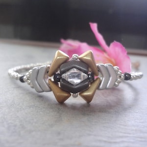 May include: A handmade bracelet featuring a central geometric design with a clear crystal, surrounded by gold, silver, and dark gray elements. The band is a delicate silver with black accents. A pink flower is in the background.