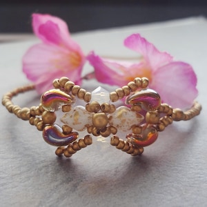 Pode incluir: Uma pulseira de contas douradas com detalhes iridescentes. A pulseira apresenta um design central com contas brancas e douradas. Flores cor-de-rosa no fundo adicionam um toque de cor e elegância à joia.