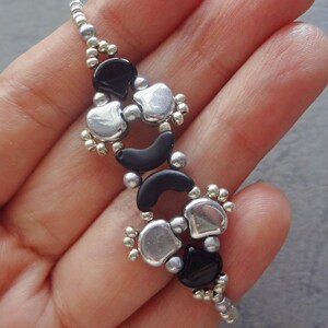 Op de afbeelding: Een handgemaakte armband met een herhalend patroon van zwarte, zilveren en kleine zilveren kralen. Het ontwerp bevat hartvormige en halvemaanvormige elementen, waardoor een uniek en elegant accessoire ontstaat. De armband wordt getoond tegen een neutrale achtergrond.