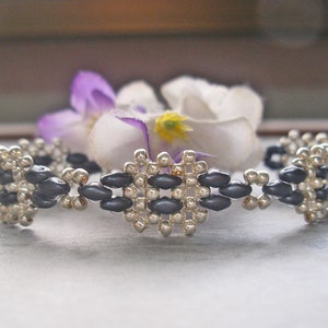 May include: A beaded bracelet featuring a central diamond-shaped design. The bracelet is made with silver and dark gray beads. The bracelet is approximately 7 inches in length.