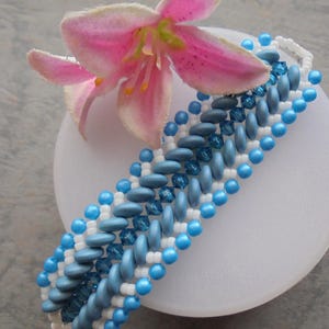 May include: A beaded bracelet featuring a pattern of light blue, dark blue, and white beads. The bracelet is adorned with small, round beads and larger, oval-shaped beads. A pink and yellow flower rests near the bracelet.