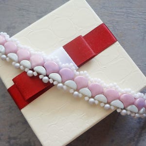 Op de afbeelding: Een kralenarmband met roze, witte en lavendelkleurige kralen, rustend op een crèmekleurige geschenkdoos met een rood lint. De armband heeft een geschulpte vormgeving met kleine, ronde kralen.
