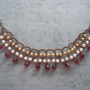 Op de afbeelding: Een kralenketting met een bronskleurige ketting, gouden en witte kralen en rode kristallen hangers. De ketting heeft een sluiting. De ketting is ongeveer 30 cm lang.