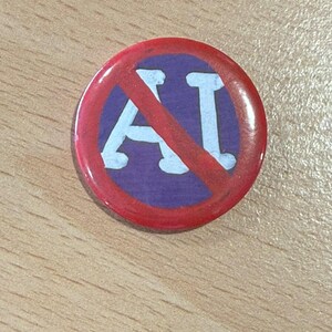 Pode incluir: Um botton circular com fundo roxo e as letras brancas AI impressas no centro. Uma linha diagonal vermelha cruza as letras, indicando um símbolo de proibição. O botton possui um acabamento brilhante e reflexivo.