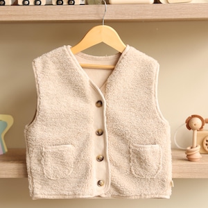 May include: A beige, fluffy fleece vest with a V-neck and four brown buttons down the front. The vest has two front pockets and is hanging on a wooden hanger. The garment is designed for children.