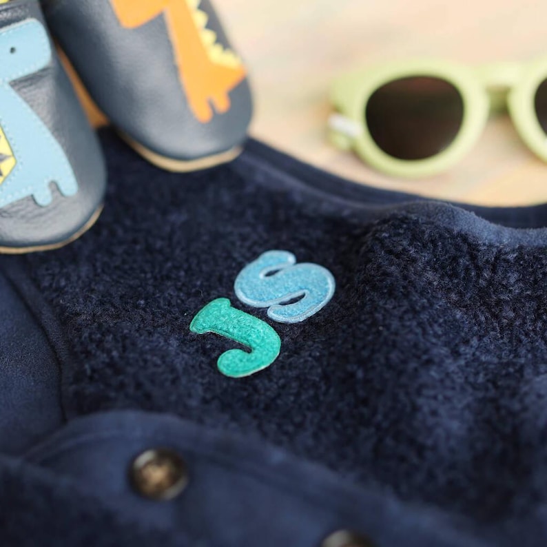May include: A navy blue baby outfit with the embroidered letters "JS" in teal and light blue. The outfit is paired with matching dinosaur-themed baby shoes and light green sunglasses.