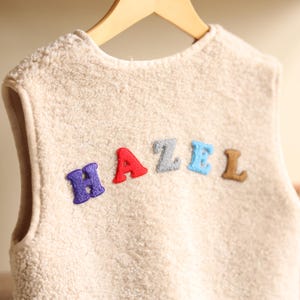 May include: A cream-colored, fuzzy vest with the name "HAZEL" embroidered across the front in colorful letters. The letters are purple, red, gray, blue, and brown. The vest is hanging on a wooden hanger.