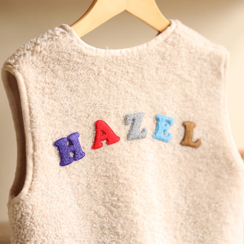 May include: A cream-colored, fuzzy vest with the name "HAZEL" embroidered across the front in colorful letters. The letters are purple, red, gray, blue, and brown. The vest is hanging on a wooden hanger.