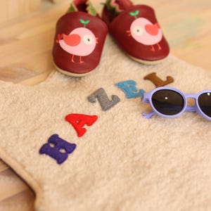 May include: A pair of burgundy leather baby shoes with a pink bird design, a beige towel with the name "HAZEL" spelled out in colorful felt letters, and a pair of purple sunglasses.