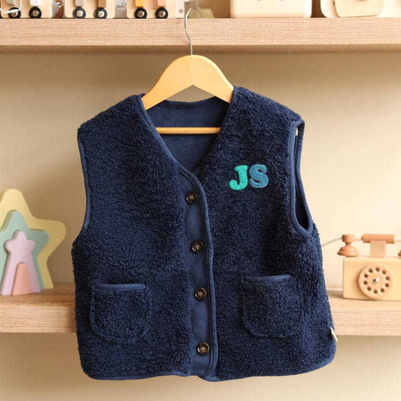 May include: A navy blue fleece vest with a V-neck, two front pockets, and wooden buttons. The vest has a patch with the letters "JS" in teal and blue. The vest is hanging on a wooden hanger.