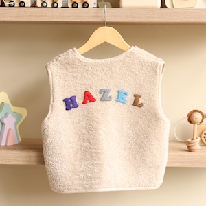 May include: A beige, sleeveless fleece vest with the name "HAZEL" embroidered across the front in colorful letters. The vest is hanging on a wooden hanger.