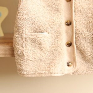 May include: Close-up of a beige, fuzzy vest with a rounded pocket and brown buttons. The vest has a soft, textured appearance, suggesting a warm and comfortable material. The buttons are evenly spaced down the front.
