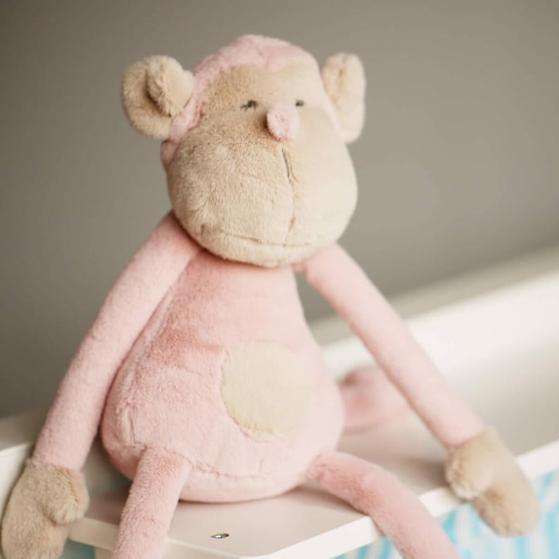 May include: A pink plush monkey toy with a beige face and a beige patch on its belly. The monkey is sitting on a white surface.