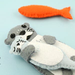May include: A handcrafted felt otter toy in shades of gray and white, holding a smaller light gray felt pup. An orange felt fish is positioned above the otters. The text Maisie Moo is printed in black script in the lower left corner of the image.