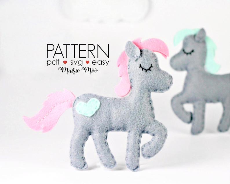 May include: A gray felt unicorn with pink mane and tail, a light blue heart on its side, and closed eyes. The unicorn is standing on a white surface. The text "PATTERN pdf &hearts; svg &hearts; easy Maisie Moo" is above the unicorn.