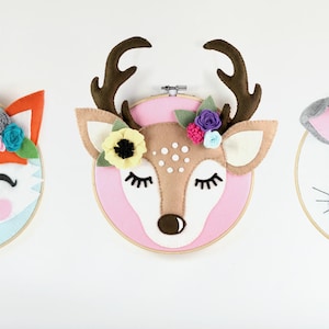 May include: A felt deer head with closed eyes and a floral crown, mounted on a pink embroidery hoop. The deer is brown with white spots and has a brown nose.